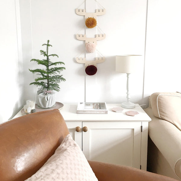 3 pompom reindeers handing on living room wall next to small christmas tree and lamp