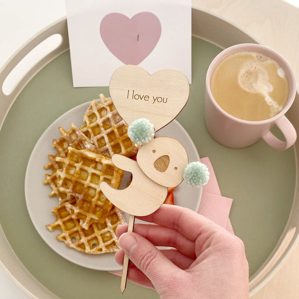 Valentines cake topper with koala and pompoms above waffles and mug of tea